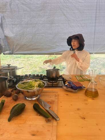 A woman in a wide-brimmed hat cooks at an outdoor kitchen, surrounded by fresh vegetables, kitchen tools, and pots under a large canopy tent.