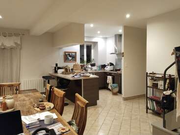 The image shows a cozy, lived-in kitchen and dining area with wooden furniture, tiled floors, scattered items on counters, shelves with books, and warm lighting.