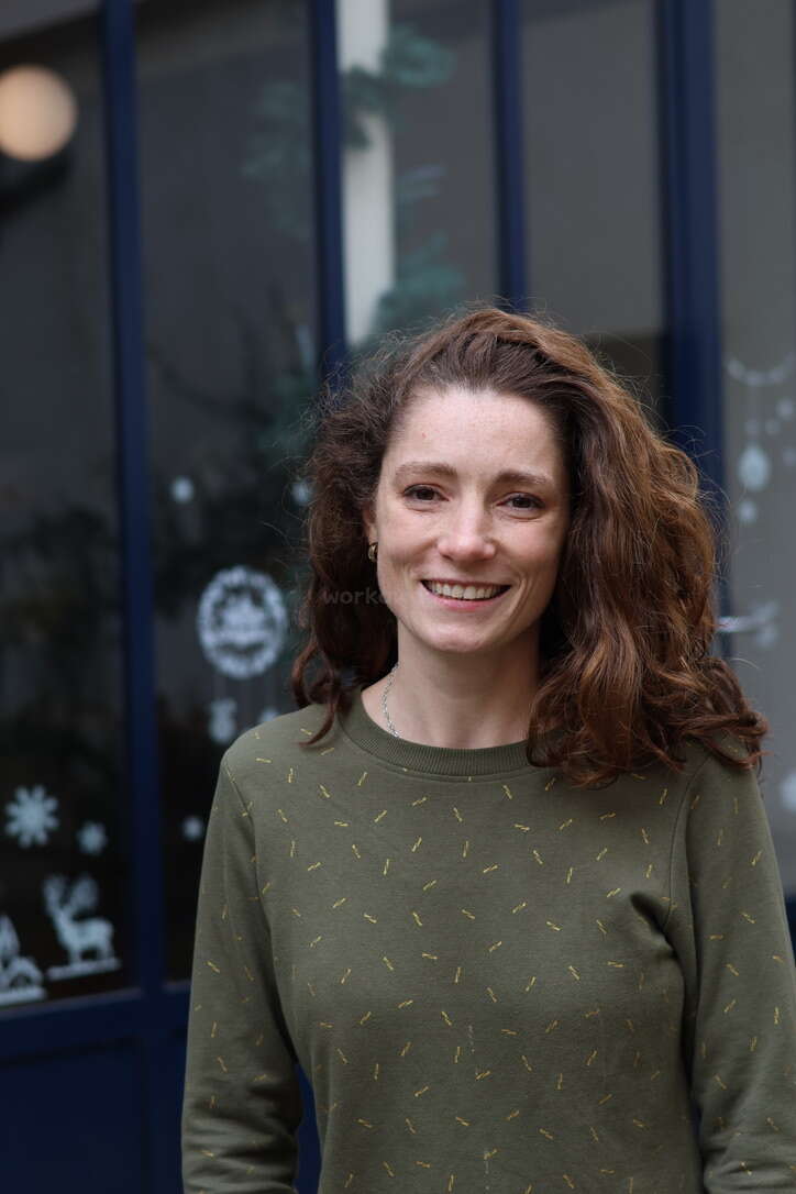 A woman with wavy brown hair smiles at the camera. She is wearing a green patterned sweater, standing in front of a window with winter decorations.