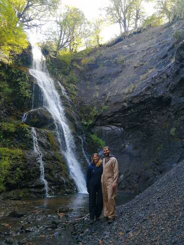 Ein lächelndes Paar steht an einem felsigen Bach am Fuße eines wunderschönen Wasserfalls, umgeben von üppigem Grün und hohen Bäumen unter einem strahlenden Himmel.