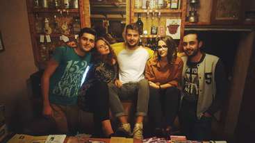 A imagem mostra um grupo de pessoas reunidas em torno de uma mesa, com alguns indivíduos em pé ao fundo. A cena parece ser uma reunião ou comemoração social.