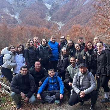 A group of people poses together outdoors in front of beautiful autumn-colored trees and mountains, dressed warmly in jackets, suggesting a hiking trip or nature outing.