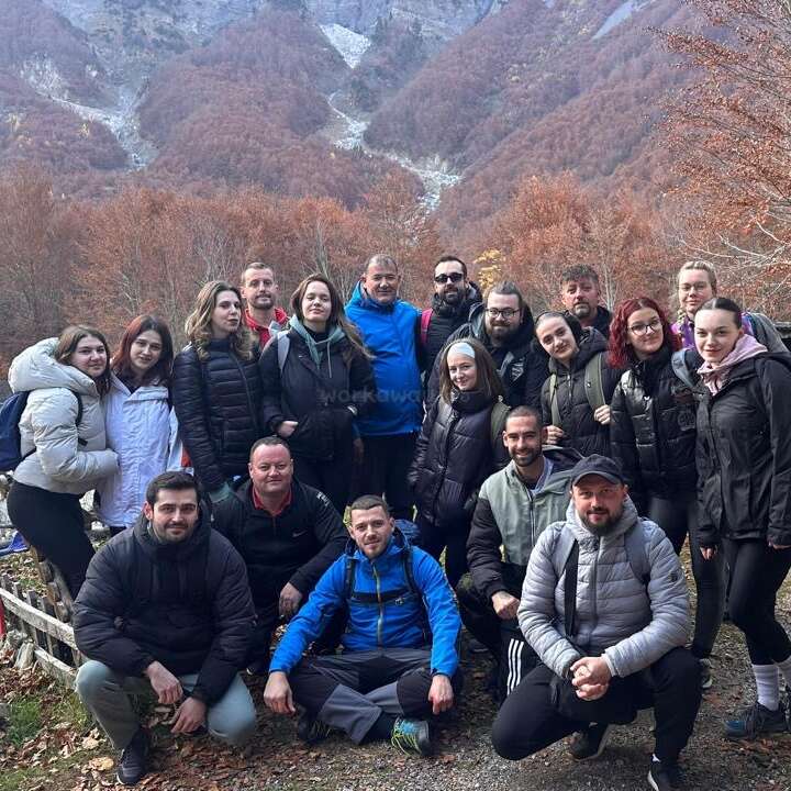 A group of people poses together outdoors in front of beautiful autumn-colored trees and mountains, dressed warmly in jackets, suggesting a hiking trip or nature outing.