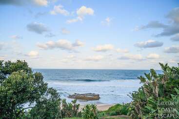 Esta imagen muestra una serena escena playera con olas azules del océano, orilla arenosa, exuberantes plantas verdes y un cielo parcialmente nublado bajo una suave luz natural.