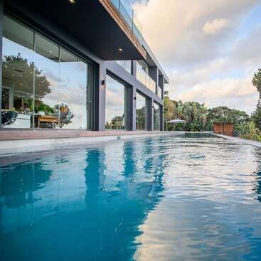 A modern house with large glass windows overlooks a serene infinity pool. Reflections shimmer in the water, surrounded by lush greenery under a cloudy sky.