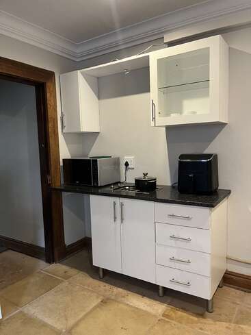 Esta imagen muestra una pequeña cocina con armarios blancos, una encimera negra, un microondas, una freidora de aire, una olla y suelo de baldosas. Un montaje sencillo y ordenado.