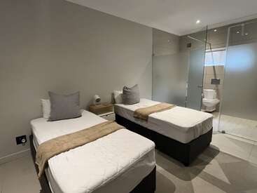 Esta imagen muestra un dormitorio moderno y minimalista con dos camas individuales, ropa de cama neutra, una mesilla de noche y un cuarto de baño acristalado con inodoro y ducha.