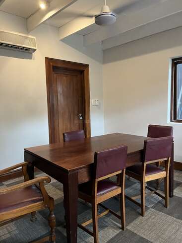 Esta imagen muestra una pequeña sala de reuniones con una mesa de madera, cuatro sillas, un aparato de aire acondicionado, un ventilador de techo, una puerta de madera y una ventana.