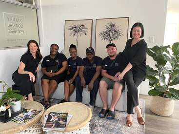 Seis personas se sientan y sonríen juntas en una sala moderna y luminosa con decoración costera, plantas de interior, revistas en las mesas y obras de arte de palmeras en la pared.