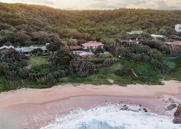 Vista aérea de serenas casas frente a la playa enclavadas entre frondosos árboles verdes, con vistas a una playa de arena con suaves olas rompiendo en la orilla bajo un cielo nublado.