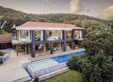 Esta imagen muestra una moderna casa de dos plantas con grandes ventanales de cristal, tejado de tejas rojas y piscina infinita, rodeada de exuberante vegetación y colinas.