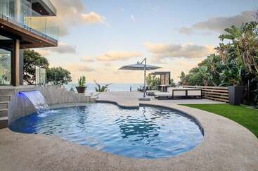 Esta imagen muestra una moderna casa de lujo con una elegante piscina, juegos de agua, muebles de jardín y un exuberante paisaje, con vistas al océano bajo un cielo sereno.