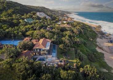Esta imagen muestra una lujosa finca frente al mar rodeada de exuberante vegetación, una piscina, una pista de tenis, varias casas y una pintoresca costa con olas rompiendo en la orilla.