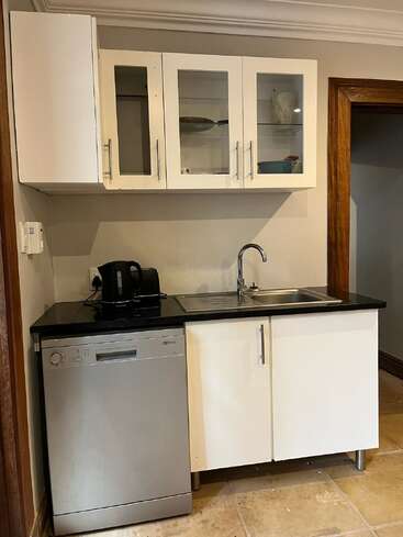 Esta imagen muestra una cocina compacta con armarios blancos, un lavavajillas de acero inoxidable, una encimera negra, un fregadero, una tetera y vitrinas encima.