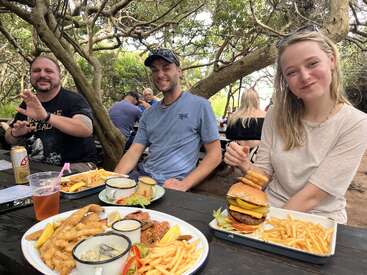 Three friends sit at an outdoor picnic table, smiling and enjoying a hearty meal of burgers, fries, fried seafood, and drinks under leafy trees in daylight.