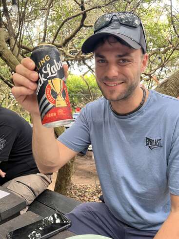 A man wearing a cap and sunglasses smiles, holding up a can of Black Label beer while sitting outdoors at a picnic table with trees in the background.