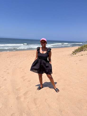 A smiling woman in a black dress and pink cap stands barefoot on a sandy beach. The sky is clear and blue, with waves in the background.