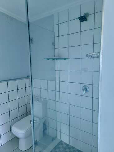 The image shows a bathroom with white square tiles, a glass shower enclosure, a modern toilet inside the shower area, a glass shelf, and a rainfall showerhead.