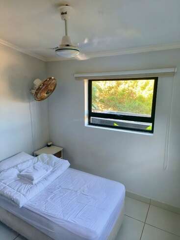 A small, clean bedroom with white walls, a single bed neatly made, ceiling fan, wall-mounted fan, side table, window with greenery outside, and tile floor.