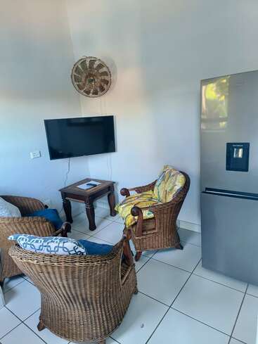 This cozy room features two wicker chairs with colorful cushions, a small wooden table, wall-mounted TV, decorative fan, and a modern refrigerator on white tiled floor.