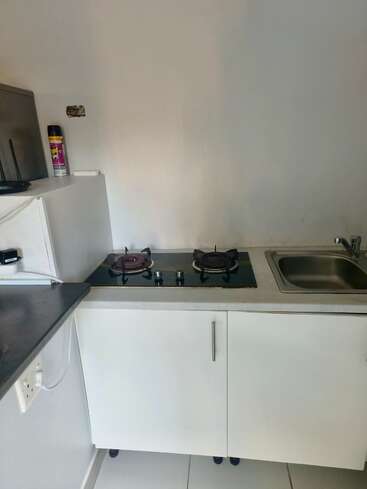 This image shows a small kitchen corner with a countertop, two-burner gas stove, stainless steel sink, white cabinets, a can of insect spray, and tiled floor.