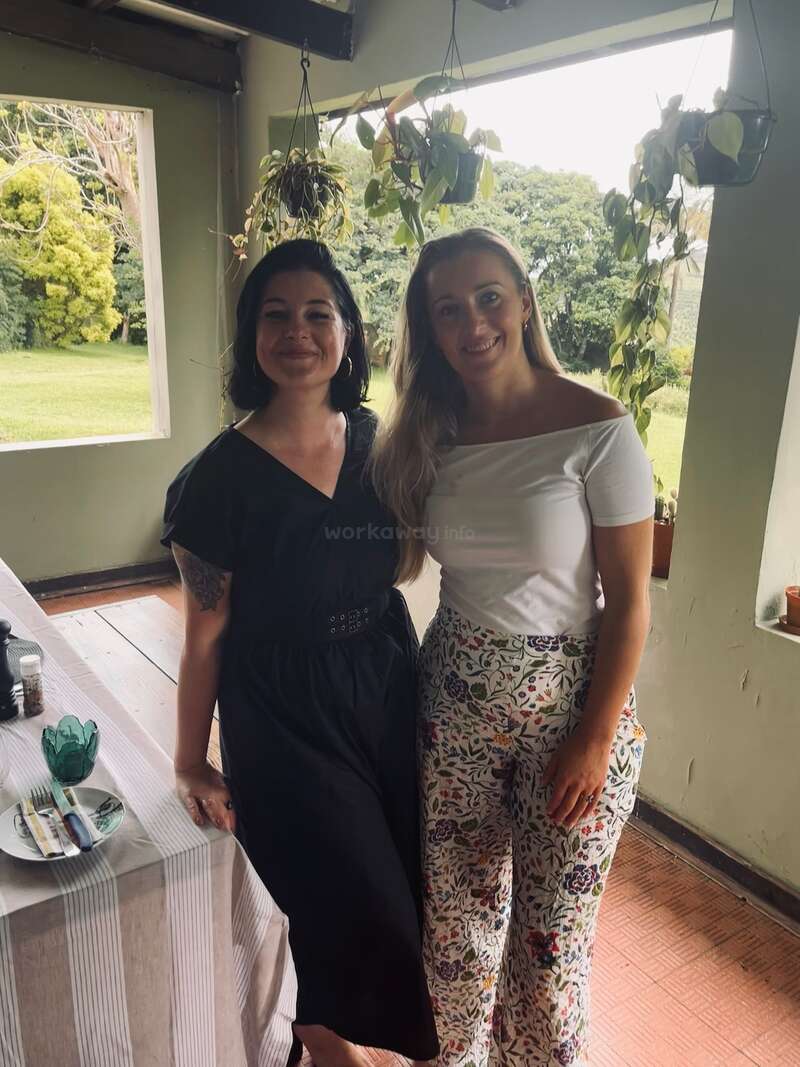 Two women are smiling and posing indoors near a window. One wears a black dress, the other a white top with floral pants. Green plants hang above.
