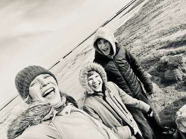 Drei Leute, die sich mit Wintermänteln und Mützen warm angezogen haben, lachen und quatschen zusammen am Strand. Das Bild ist schwarz-weiß und fängt fröhliche Momente ein.