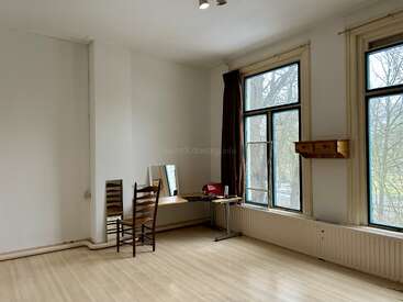 Ein geräumiges, minimalistisches Zimmer mit großen Fenstern, hellen Holzböden, zwei Stühlen, einem einfachen Schreibtisch, einem Spiegel, ein paar kleinen Sachen und Blick auf die Bäume draußen.