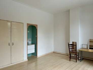 Auf dem Bild sieht man einen fast leeren Raum mit hellen Holzböden, weißen Wänden, einer kleinen Spüle, einem Schrank, einem Holzstuhl und einem einfachen Schreibtisch.