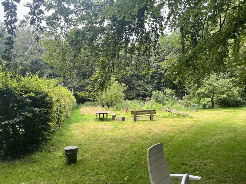 Das Bild zeigt einen ruhigen, grünen Garten mit Bänken, Tischen und ein paar Gartenmöbeln. Hohe Bäume, eine Hecke und hohes Gras sorgen für eine entspannte, natürliche Atmosphäre.