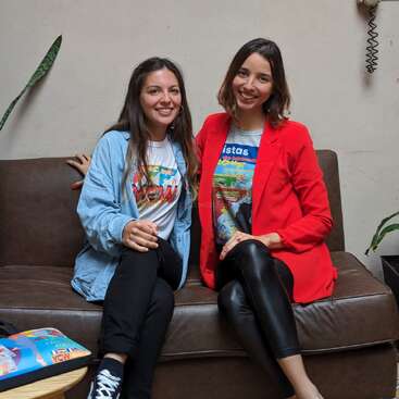 Duas mulheres sentadas em um sofá de couro marrom, vestindo roupas casuais e sorrindo para a câmera, com uma planta ao fundo e uma mesa com uma bolsa colorida à sua esquerda.