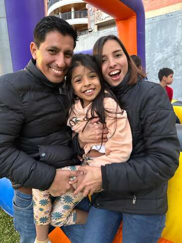 Das Bild zeigt eine dreiköpfige Familie, die für ein Foto vor einer aufblasbaren Hüpfburg posiert, während die Eltern ihre kleine Tochter umarmen.