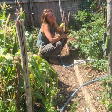 Una mujer cuida de un jardín, de pie detrás de una hilera de plantas, con una valla y árboles visibles al fondo, rodeada de verdor y follaje.
