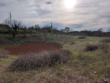 A cloudy sky covers a rural landscape with a plowed circular field, dry grass, scattered bushes, leafless trees, and distant power lines in the background.