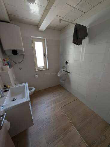 This is a modern bathroom with wood-style flooring, white tiles, a walk-in shower, a sink, toilet, water heater, window, and dark towels hanging.