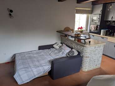 A cozy studio apartment with a pull-out sofa bed, gray bedding, brick kitchen island, modern kitchen, wooden beams, natural light, and minimalistic decor featuring throw pillows.
