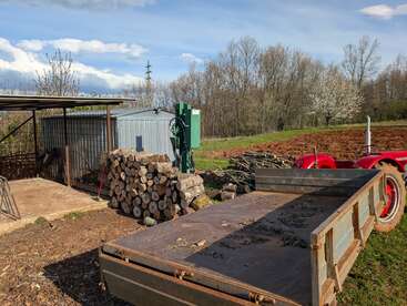 A rural scene with stacked firewood, a shed, a metal machine, a field, a red tractor, trailer, some trees, and a clear, partly cloudy sky.