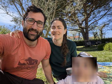 A man, woman, and a child (face blurred) enjoy a sunny day outdoors, relaxing on grass with trees, blue sky, and a swing in the background.