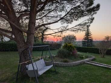 A peaceful garden at sunset, featuring a large tree, a cozy swing, flower beds, manicured grass, distant shrubs, and a glowing orange and pink sky.