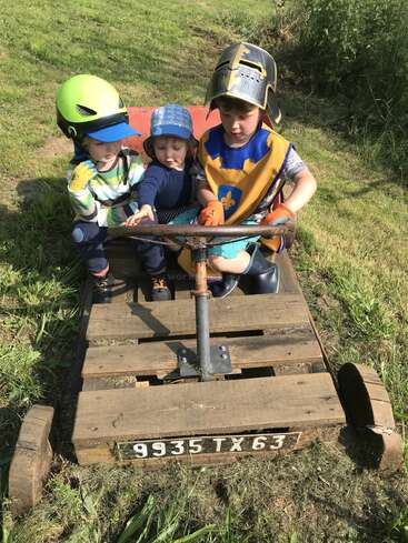 Drei Kinder sitzen auf einem selbstgebauten Go-Kart aus Holz auf einer Wiese. Sie tragen verspielte Kostüme und Helme, lächeln und genießen ihr gemeinsames fantasievolles Abenteuer im Freien.