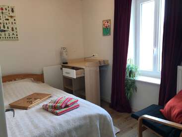 Dieses gemütliche Schlafzimmer verfügt über ein Bett mit gefalteten Handtüchern, einen Schreibtisch, einen Stuhl, eine Zimmerpflanze, rote Vorhänge, ein Insektenkunstwerk, ein helles Fenster und einen Holzfußboden.