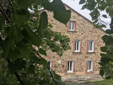 Das Bild zeigt ein steinernes Gebäude mit mehreren Fenstern, durch grüne Blätter betrachtet, vor einem bedeckten Himmel. Die Fassade des Gebäudes ist aus hellbraunen und braunen Steinen gebaut.