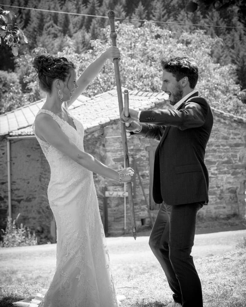 Das Bild zeigt eine Braut und einen Bräutigam, die zusammen stehen und eine lange Stange mit einer dekorativen Spitze halten, vor einem rustikalen Steinhaus und Bäumen im Hintergrund.