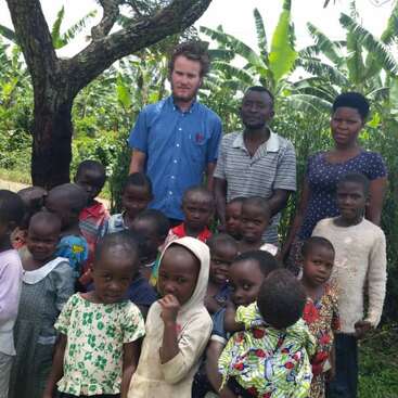 A group of children and three adults stand together outdoors under a tree, surrounded by green vegetation. The children wear colorful clothing, looking towards the camera.