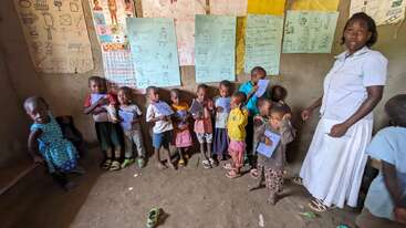 Un grupo de niños pequeños está de pie en un aula, sosteniendo papeles. La pared está cubierta de carteles coloridos y materiales didácticos. Una profesora está de pie cerca de ellos, observándolos.