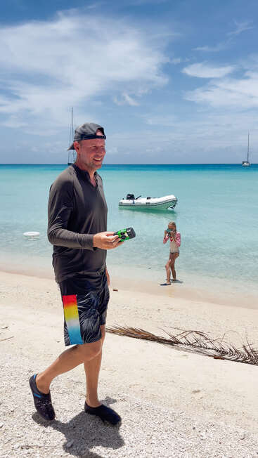 Das Bild zeigt einen Mann, der an einem Strand spazieren geht und ein Spielzeugauto in der Hand hält, während im Hintergrund ein Kind steht und dahinter Boote im Wasser treiben.