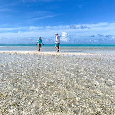 Das Bild zeigt eine ruhige Strandszene mit zwei Personen, die am Ufer entlang spazieren, umgeben von kristallklarem Wasser und einem strahlend blauen Himmel.