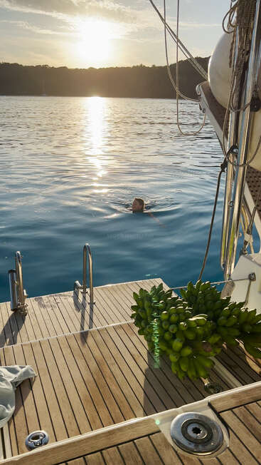 Das Bild zeigt eine heitere Szene, in der eine Person vom Deck eines Bootes aus im Wasser schwimmt, mit einem großen Bündel Bananen auf dem Deck und einem Sonnenuntergang im Hintergrund.
