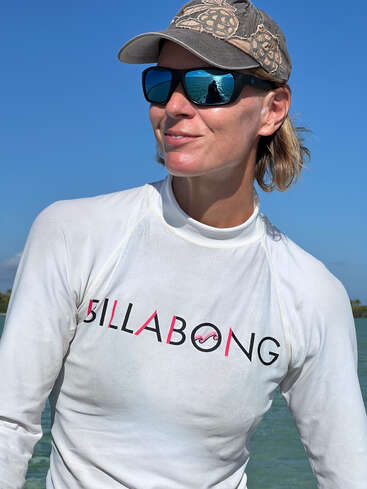 Das Bild zeigt eine Frau, die vor einem Gewässer steht und ein weißes Billabong-Shirt, eine Sonnenbrille und eine Baseballmütze in Tarnfarben trägt.