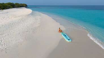Eine Person sitzt an einem unberührten Sandstrand neben einem Paddleboard, umgeben von türkisblauem Wasser und klarem Himmel, mit üppig grünen Bäumen in der Ferne.